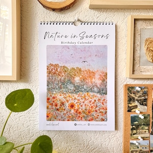 May include: A spiral-bound birthday calendar titled Nature in Seasons featuring a watercolour-style illustration of a field of orange and yellow flowers under a sky with birds. The calendar includes the text anli liu art and a website address.