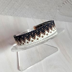 Könnte beinhalten: Ein Armband mit Perlen und einem geometrischen Muster. Das Armband hat eine Kombination aus marineblauen, weißen und kupferfarbenen Perlen. Das Armband wird auf einem transparenten, abgewinkelten Ständer präsentiert, der sein Design und seine Handwerkskunst zeigt. Es hat einen Metallverschluss.