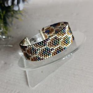 May include: A wide beaded bracelet with a silver clasp. The bracelet features a pattern of small, colorful beads in shades of gold, brown, blue, and white. The bracelet is displayed on a clear, angled stand.