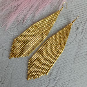 Op de afbeelding: Een paar gouden kralen oorbellen met een franje ontwerp. De oorbellen hebben een driehoekige vorm, met lange, verticale strengen gouden kralen. De haken zijn goudkleurig. De achtergrond is een lichtgrijze ondergrond met een vleugje roze.