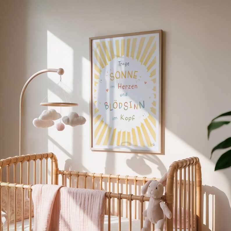 May include: A framed print with the text "Trage Sonne im Herzen und Bl&ouml;dsinn im Kopf" in a yellow sunburst design. A baby crib with a pink blanket and a stuffed bunny is in the foreground. A mobile with clouds and a pink ball hangs above the crib.