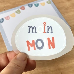 May include: A white sticker with a stitched border and the text "moin MOIN" in black, red, and white lettering. The sticker is on a blue and white patterned background.