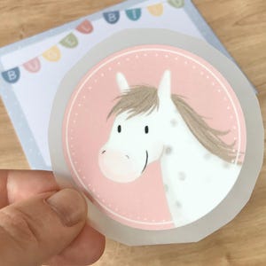Puede incluir: Un caballo blanco con pelo marrón y ojos marrones sobre un fondo rosa con lunares blancos. El caballo está mirando hacia la izquierda. La imagen está en un círculo transparente con un borde blanco.