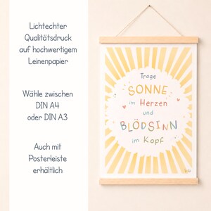 May include: A framed art print with a wooden hanger. The print features a yellow sunburst design with the words "SONNE im Herzen und BL&Ouml;DSINN im Kopf" in colorful lettering. Additional text in German is also present.