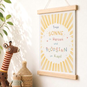 May include: A framed print with a wooden hanger displays the text "Trage Sonne im Herzen und Bl&ouml;dsinn im Kopf" in colorful lettering. The background features a yellow sunburst design. A crocheted giraffe and other toys are in the foreground.
