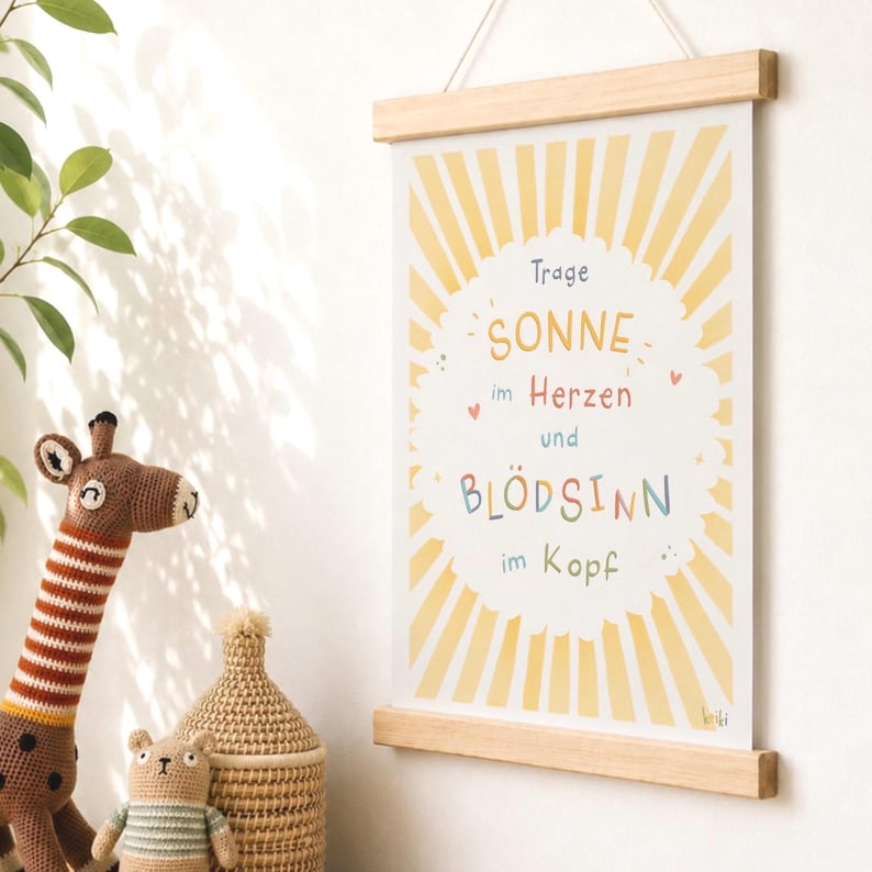 May include: A framed print with a wooden hanger displays the text "Trage Sonne im Herzen und Bl&ouml;dsinn im Kopf" in colorful lettering. The background features a yellow sunburst design. A crocheted giraffe and other toys are in the foreground.