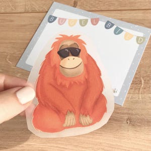 Puede incluir: Un orangután de color naranja brillante con gafas de sol, sentado con las piernas cruzadas. El orangután está sobre un fondo blanco con la palabra "Bügelbild" en letras de colores en la parte superior.