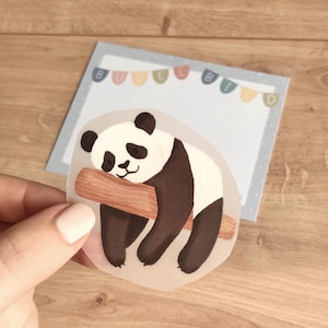 May include: A sticker featuring a sleeping panda bear with black and white fur, resting on a brown log. The sticker is held in front of a light blue notepad with the text "BUBBLE BIRD" on top.