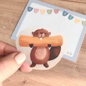 Könnte beinhalten: Ein brauner Cartoon-Biber mit einem zahnigen Grinsen hält einen großen, hellbraunen Baumstamm in seinen Pfoten. Der Biber befindet sich auf einem durchsichtigen Aufkleber mit weißem Hintergrund.