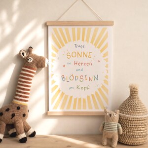 May include: A framed poster with the text "SONNE im Herzen und BL&Ouml;DSINN im Kopf" in colorful letters, hanging above a wooden surface. A crocheted giraffe and a small bear are on the surface, along with a woven basket.