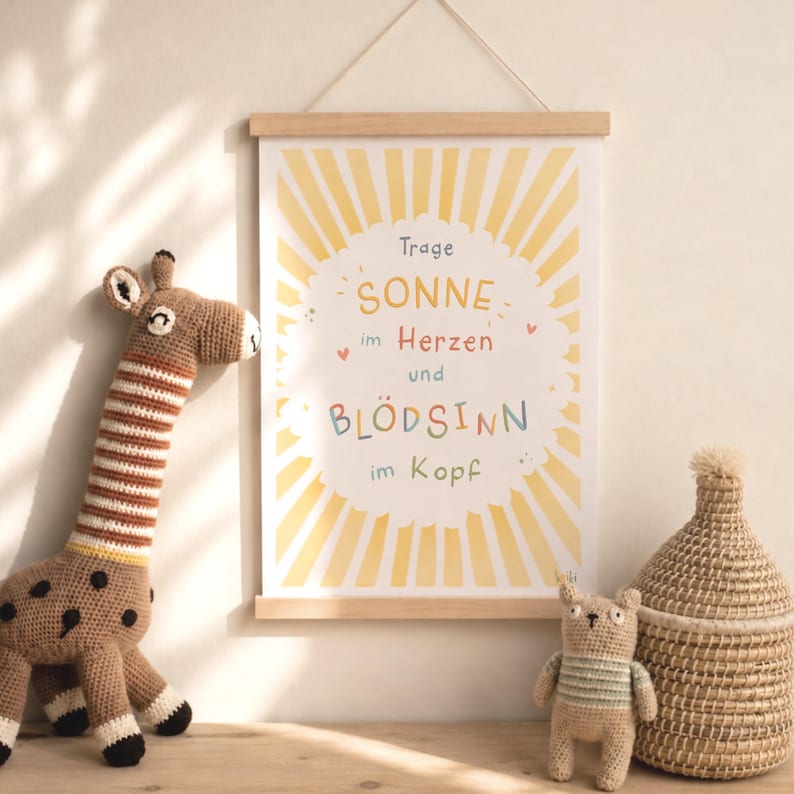 May include: A framed poster with the text "SONNE im Herzen und BL&Ouml;DSINN im Kopf" in colorful letters, hanging above a wooden surface. A crocheted giraffe and a small bear are on the surface, along with a woven basket.