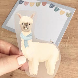 May include: A white llama with a blue scarf, illustrated on a clear sticker. The sticker is on a light blue background with the text "Bügelbild" in white letters.