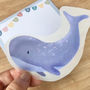 Puede incluir: Una pegatina de ballena azul con fondo blanco. La ballena tiene una barriga azul claro y una franja blanca que recorre su costado. La ballena está rodeada por un borde blanco.