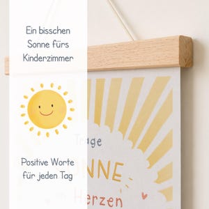 May include: A cheerful, framed print with a smiling yellow sun illustration and the words "Positive Words for Every Day." The print is held by a natural wood frame and a string for hanging. The text "Ein bisschen Sonne f&uuml;rs Kinderzimmer" is also visible.