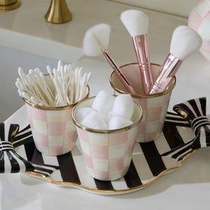 May include: A decorative tray with black and white stripes and bow-shaped handles, holding three matching ceramic cups with a pink and white checkered pattern. The cups contain white cotton swabs, white cotton balls, and makeup brushes.