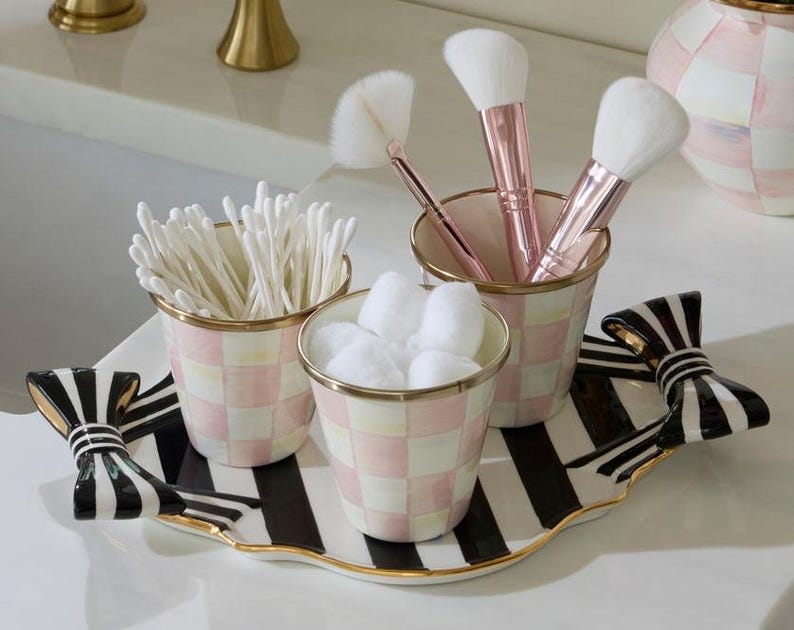 May include: A decorative tray with black and white stripes and bow-shaped handles, holding three matching ceramic cups with a pink and white checkered pattern. The cups contain white cotton swabs, white cotton balls, and makeup brushes.