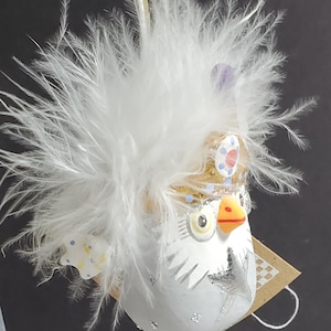 May include: A whimsical owl ornament with a white body, adorned with a fluffy white feather headdress. The owl features a gold crown, blue and yellow accents, and a silver star design. A charming decorative piece.