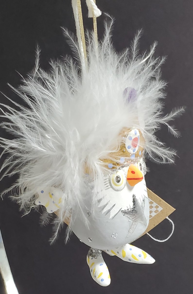 May include: A whimsical owl ornament with a white body, adorned with a fluffy white feather headdress. The owl features a gold crown, blue and yellow accents, and a silver star design. A charming decorative piece.