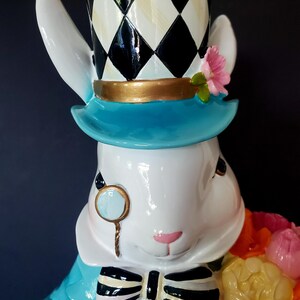 Puede incluir: Figura de conejo blanco con chaqueta azul, sombrero de copa a cuadros blanco y negro y ala azul. El conejo lleva un mon&oacute;culo con borde dorado y una pajarita en blanco y negro. Sostiene un ramo de flores coloridas.