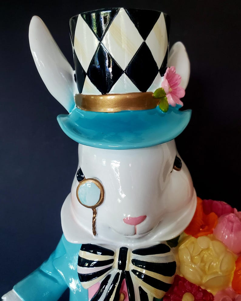 Puede incluir: Figura de conejo blanco con chaqueta azul, sombrero de copa a cuadros blanco y negro y ala azul. El conejo lleva un mon&oacute;culo con borde dorado y una pajarita en blanco y negro. Sostiene un ramo de flores coloridas.