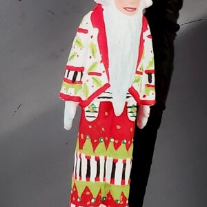 May include: A whimsical Christmas ornament depicting a figure with a crown and long white beard. The figure wears a red, white, and green patterned coat and skirt, with gold shoes. A gold string is attached to the ornament.