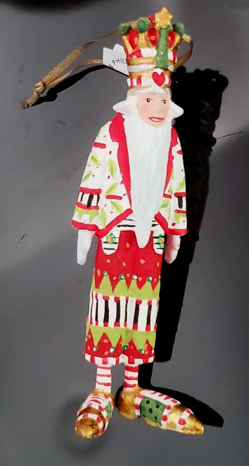 May include: A whimsical Christmas ornament depicting a figure with a crown and long white beard. The figure wears a red, white, and green patterned coat and skirt, with gold shoes. A gold string is attached to the ornament.