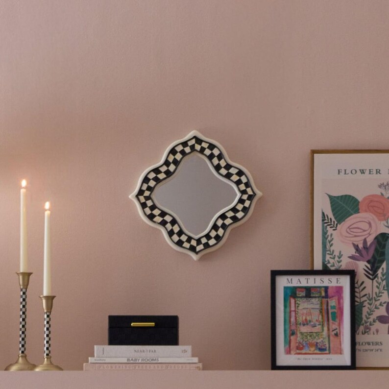 Puede incluir: Un espejo de pared con forma de cuatrifolio y un borde a cuadros blancos y negros. El espejo est&aacute; colocado sobre una pila de libros, una caja negra, dos candelabros altos con tallos a cuadros y dos l&aacute;minas de arte enmarcadas en una pared.
