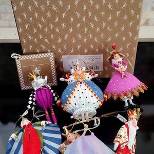 May include: A collection of decorative ornaments featuring whimsical characters in vibrant colors. The ornaments include a queen, a king, and other figures with detailed designs. They are displayed in front of a patterned box.