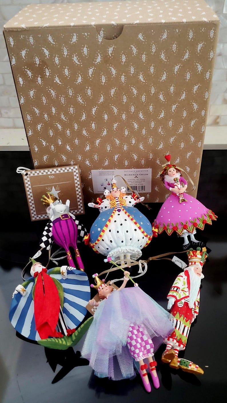 May include: A collection of decorative ornaments featuring whimsical characters in vibrant colors. The ornaments include a queen, a king, and other figures with detailed designs. They are displayed in front of a patterned box.