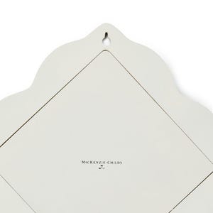 Puede incluir: La parte posterior de un objeto de pared de color crema con un borde superior festoneado y un colgador central en forma de cerradura. La superficie es lisa y presenta el texto negro MacKenzie-Childs impreso en el centro, indicando la marca.