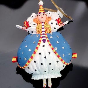 May include: A whimsical Christmas ornament shaped like a queen, featuring a blue dress with silver accents and black polka dots. The ornament has a white ruffled skirt, a red, yellow, and white striped bodice, and a gold hanging ribbon.