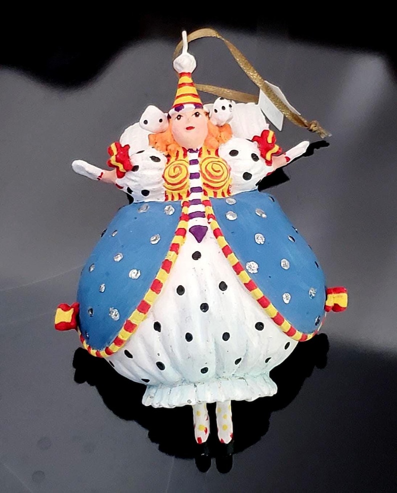 May include: A whimsical Christmas ornament shaped like a queen, featuring a blue dress with silver accents and black polka dots. The ornament has a white ruffled skirt, a red, yellow, and white striped bodice, and a gold hanging ribbon.
