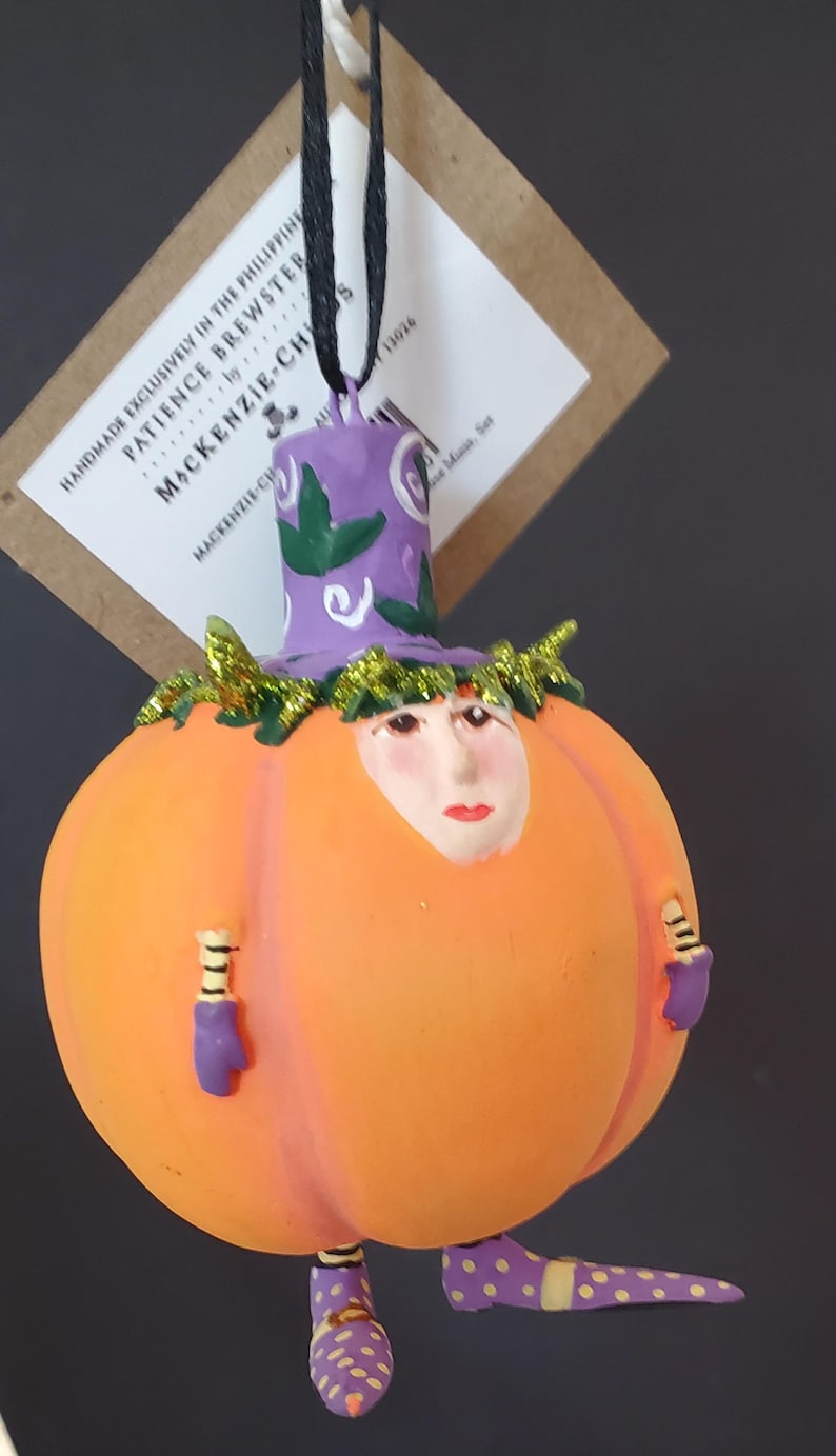 May include: A quirky Halloween ornament with a pumpkin body, painted face, purple hat and shoes. The ornament has small arms and legs, decorated with glitter and polka dots. The tag reads "MACKENZIE-CH".