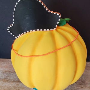 May include: A decorative, yellow pumpkin ornament with a black pirate hat. The hat features a white and black striped trim. An orange string wraps around the pumpkin. A green leaf is visible. The base has a colourful striped pattern.