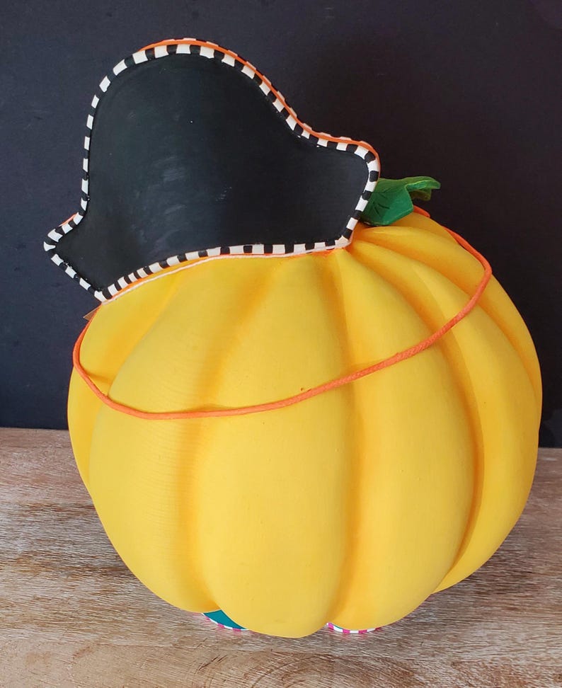 May include: A decorative, yellow pumpkin ornament with a black pirate hat. The hat features a white and black striped trim. An orange string wraps around the pumpkin. A green leaf is visible. The base has a colourful striped pattern.