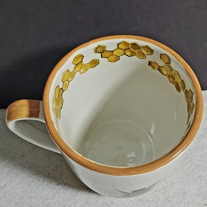May include: A white ceramic mug featuring a hand-painted golden yellow honeycomb pattern along the interior rim. The mug has a smooth, glossy finish, a rounded handle, and a solid light brown painted edge around the top opening of the vessel.