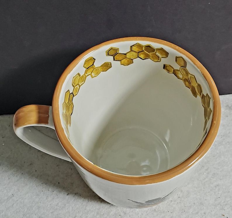 May include: A white ceramic mug featuring a hand-painted golden yellow honeycomb pattern along the interior rim. The mug has a smooth, glossy finish, a rounded handle, and a solid light brown painted edge around the top opening of the vessel.