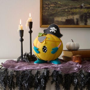May include: A novelty Halloween pumpkin ornament styled as a pirate, with a black hat and eye patch. The yellow pumpkin has blue shoes and arms. Two lit candles in black holders are positioned to the left of the pumpkin.