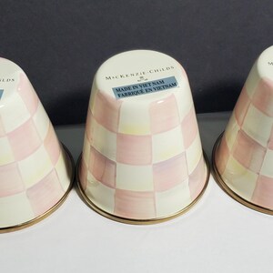 May include: Three inverted ceramic tumblers featuring a light pink and cream checkered pattern. Each tumbler has a gold-tone rim at the base and a label near the top reading MacKenzie-Childs and Made in Viet Nam Fabrique en Vietnam.
