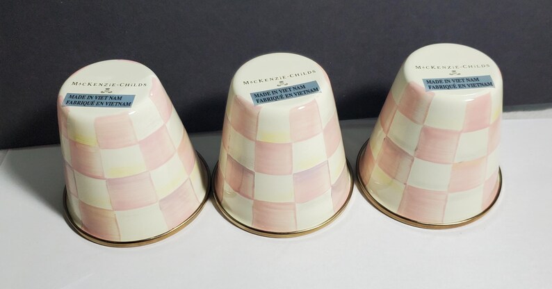 May include: Three inverted ceramic tumblers featuring a light pink and cream checkered pattern. Each tumbler has a gold-tone rim at the base and a label near the top reading MacKenzie-Childs and Made in Viet Nam Fabrique en Vietnam.