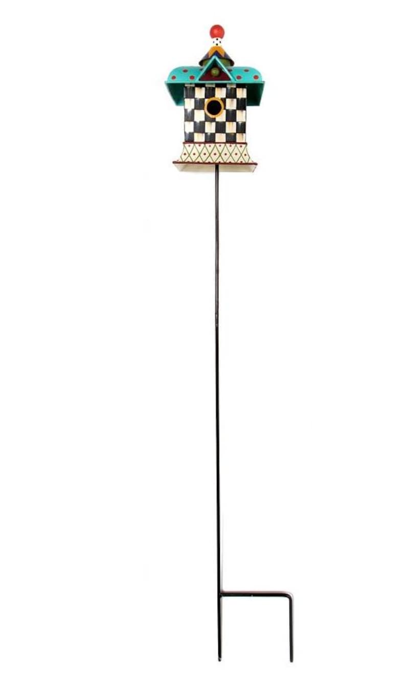 May include: A decorative birdhouse on a tall black metal garden stake. The birdhouse features a black and white checkered body, a teal roof with polka dots, and a small circular entrance hole. A red sphere sits atop the pointed roof structure.