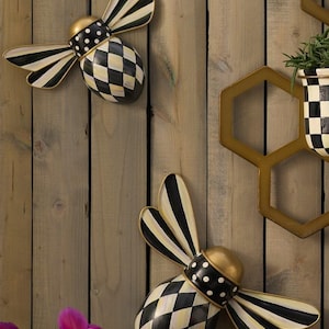Puede incluir: Dos adornos de pared decorativos con forma de abeja, con alas a rayas blancas y negras, cabeza dorada, cuello con lunares blancos y negros y cuerpo con patr&oacute;n de rombos blancos y negros. Un marco hexagonal dorado es visible a la derecha.