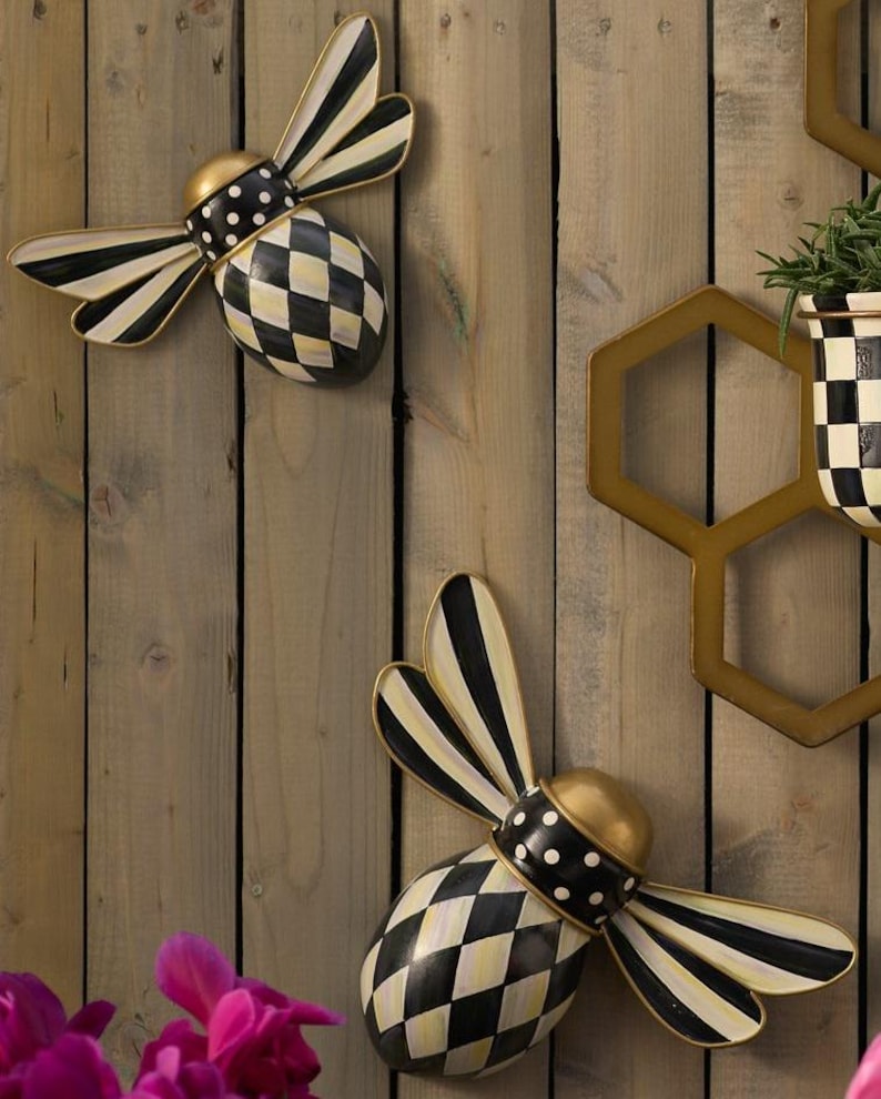Puede incluir: Dos adornos de pared decorativos con forma de abeja, con alas a rayas blancas y negras, cabeza dorada, cuello con lunares blancos y negros y cuerpo con patr&oacute;n de rombos blancos y negros. Un marco hexagonal dorado es visible a la derecha.