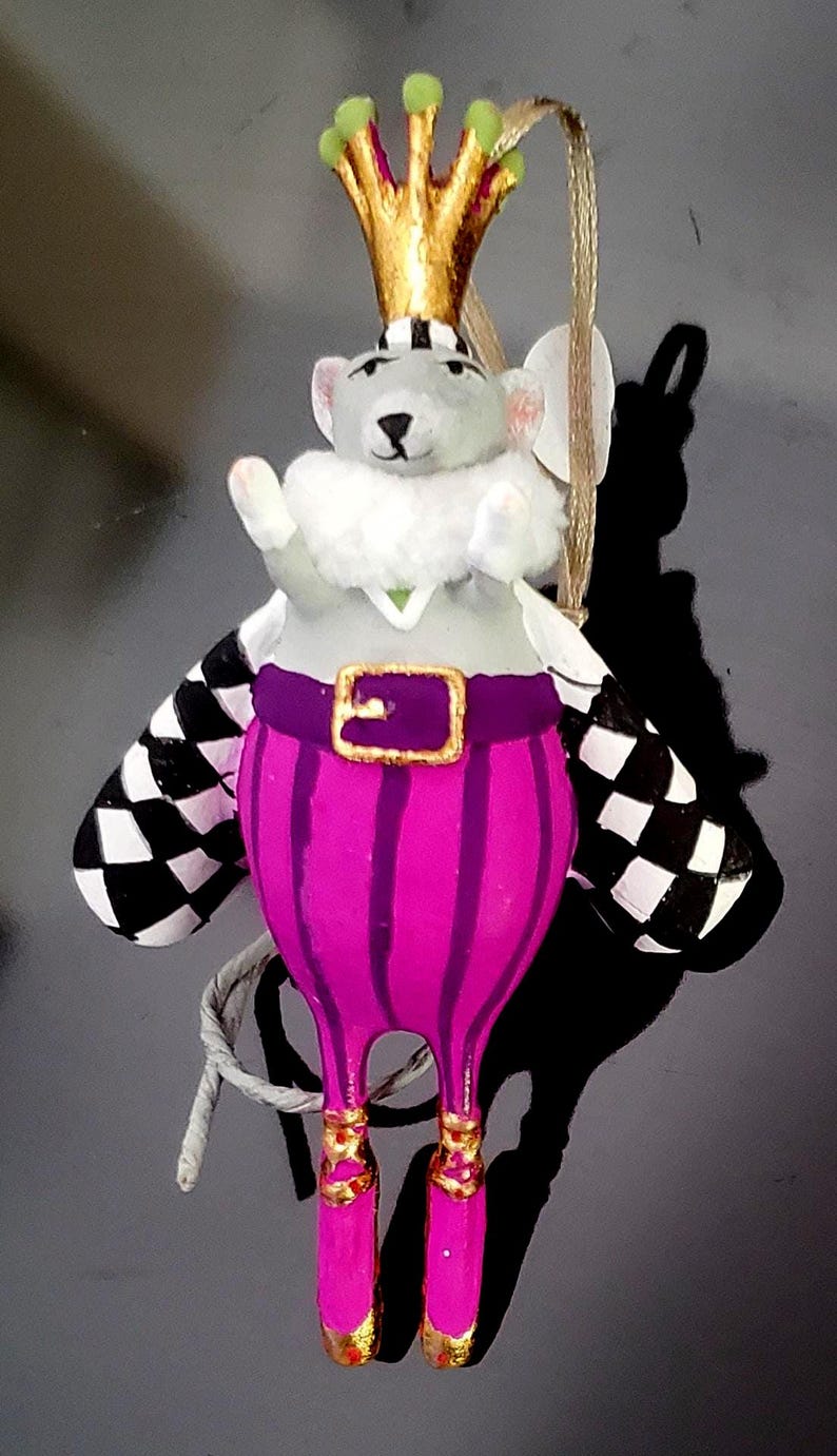 May include: A whimsical ornament of a mouse dressed as a king. The mouse has a gold crown, a white fur collar, and a purple outfit with gold accents. The mouse has black and white checkered wings and pink shoes. A gold string is attached for hanging.