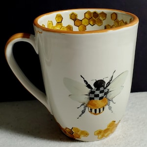 May include: A white ceramic mug featuring a hand-painted bee with a black and white checkered thorax and striped abdomen. The mug is decorated with yellow honeycomb patterns around the base and along the interior rim, finished with an orange rim.