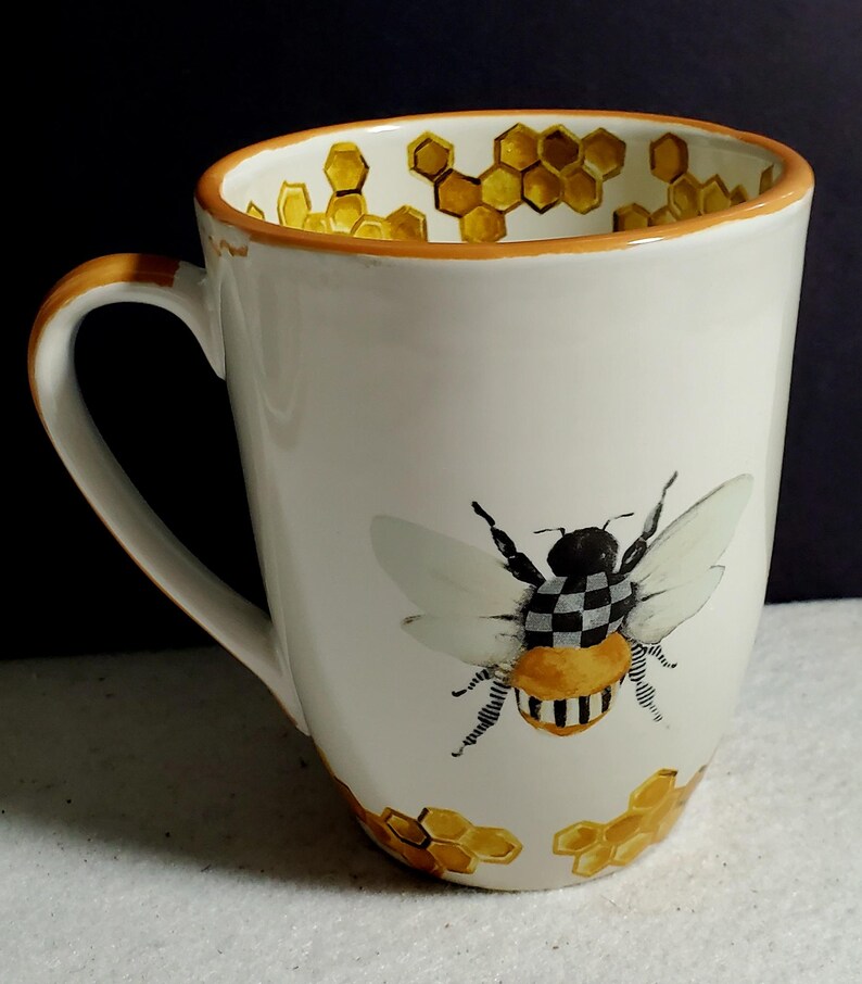 May include: A white ceramic mug featuring a hand-painted bee with a black and white checkered thorax and striped abdomen. The mug is decorated with yellow honeycomb patterns around the base and along the interior rim, finished with an orange rim.
