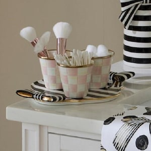 May include: A vanity set featuring three small cups with a pink and white checkered pattern and gold rims, resting on an oval tray with black and white stripes and gold bow accents. The cups hold makeup brushes and cotton swabs.