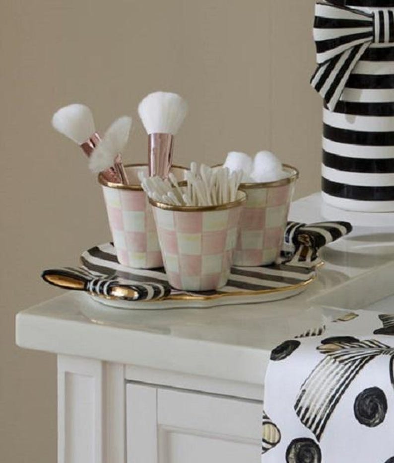 May include: A vanity set featuring three small cups with a pink and white checkered pattern and gold rims, resting on an oval tray with black and white stripes and gold bow accents. The cups hold makeup brushes and cotton swabs.
