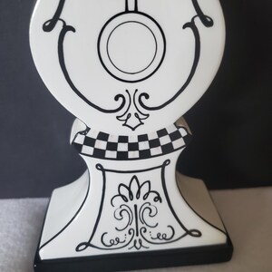 May include: A decorative white clock figurine with black accents. The clock features a pendulum design, a circular clock face, and ornate scrollwork. The base has a black and white checkered pattern and a black border.