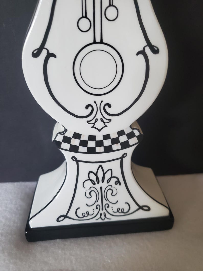 May include: A decorative white clock figurine with black accents. The clock features a pendulum design, a circular clock face, and ornate scrollwork. The base has a black and white checkered pattern and a black border.